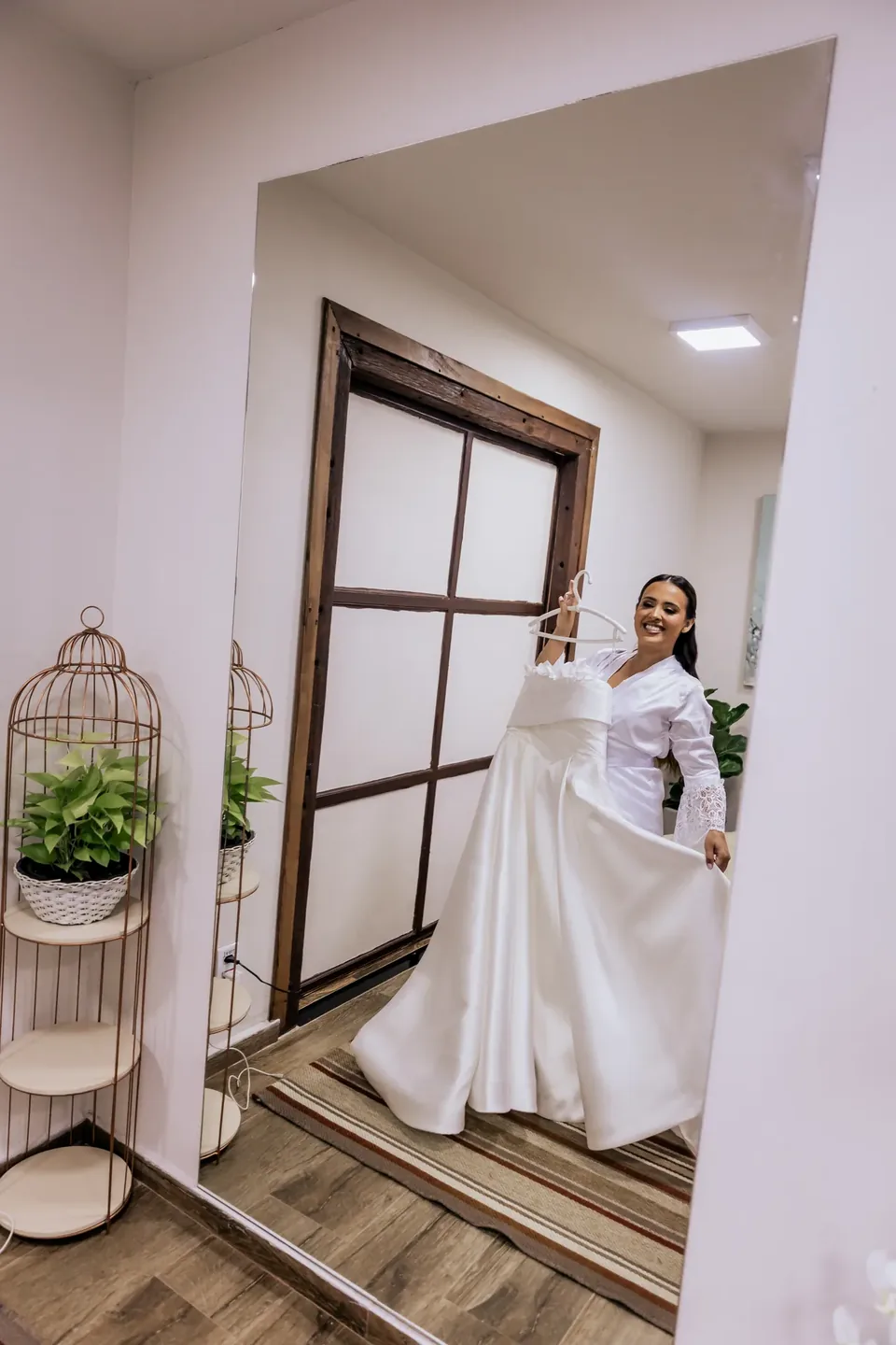 Noiva segurando o vestido no cabide em frente à janela de madeira do camarim