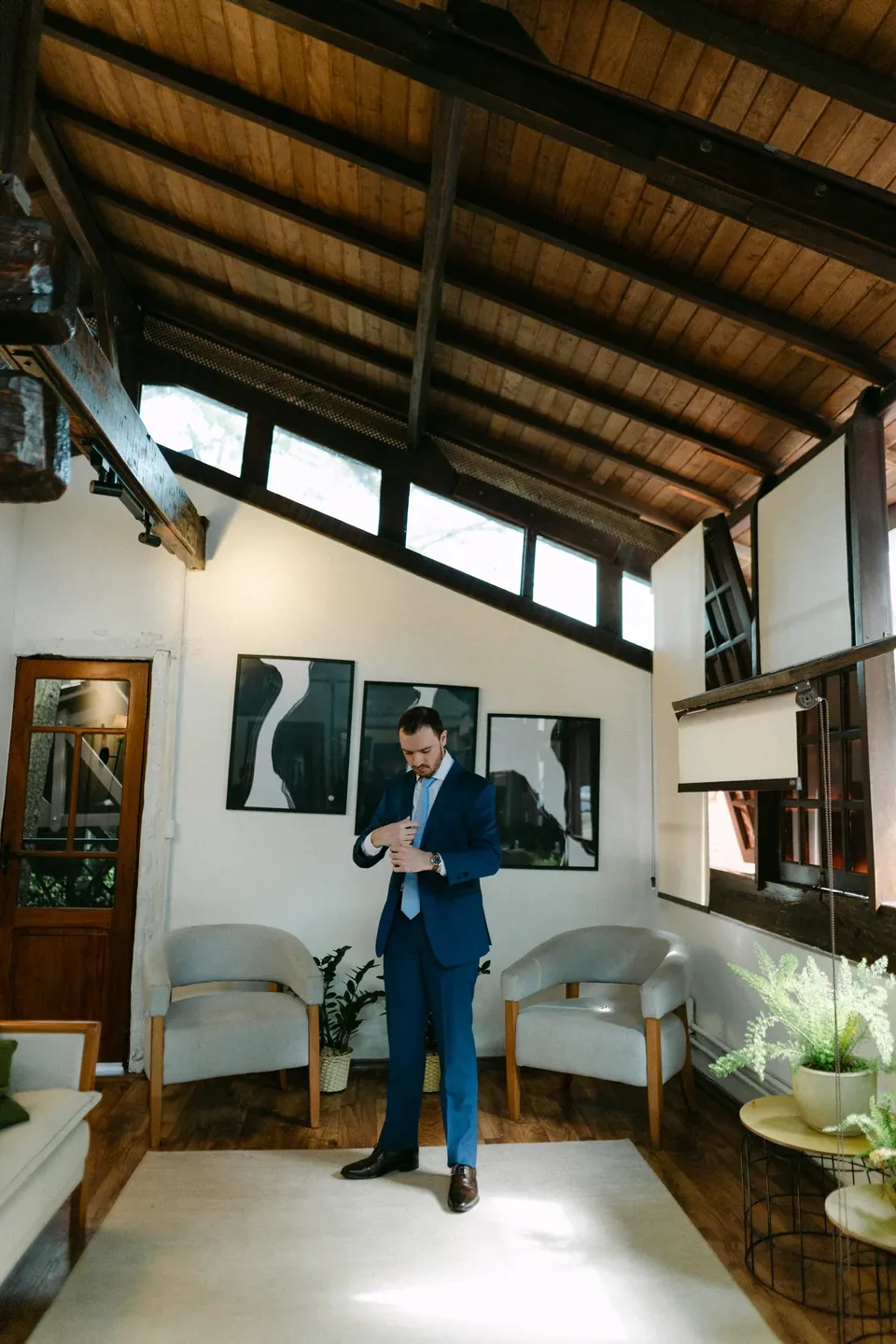 Noivo de terno azul ajustando o punho da camisa na sala do camarim, sob o telhado de madeira