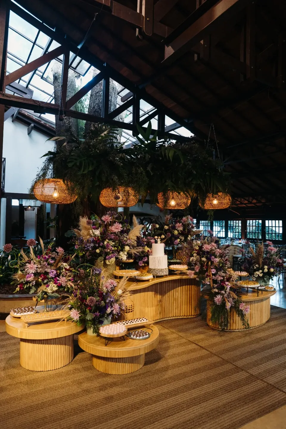 Mesa de bolo e doces com flores em tons de lilás e luminárias de fibra