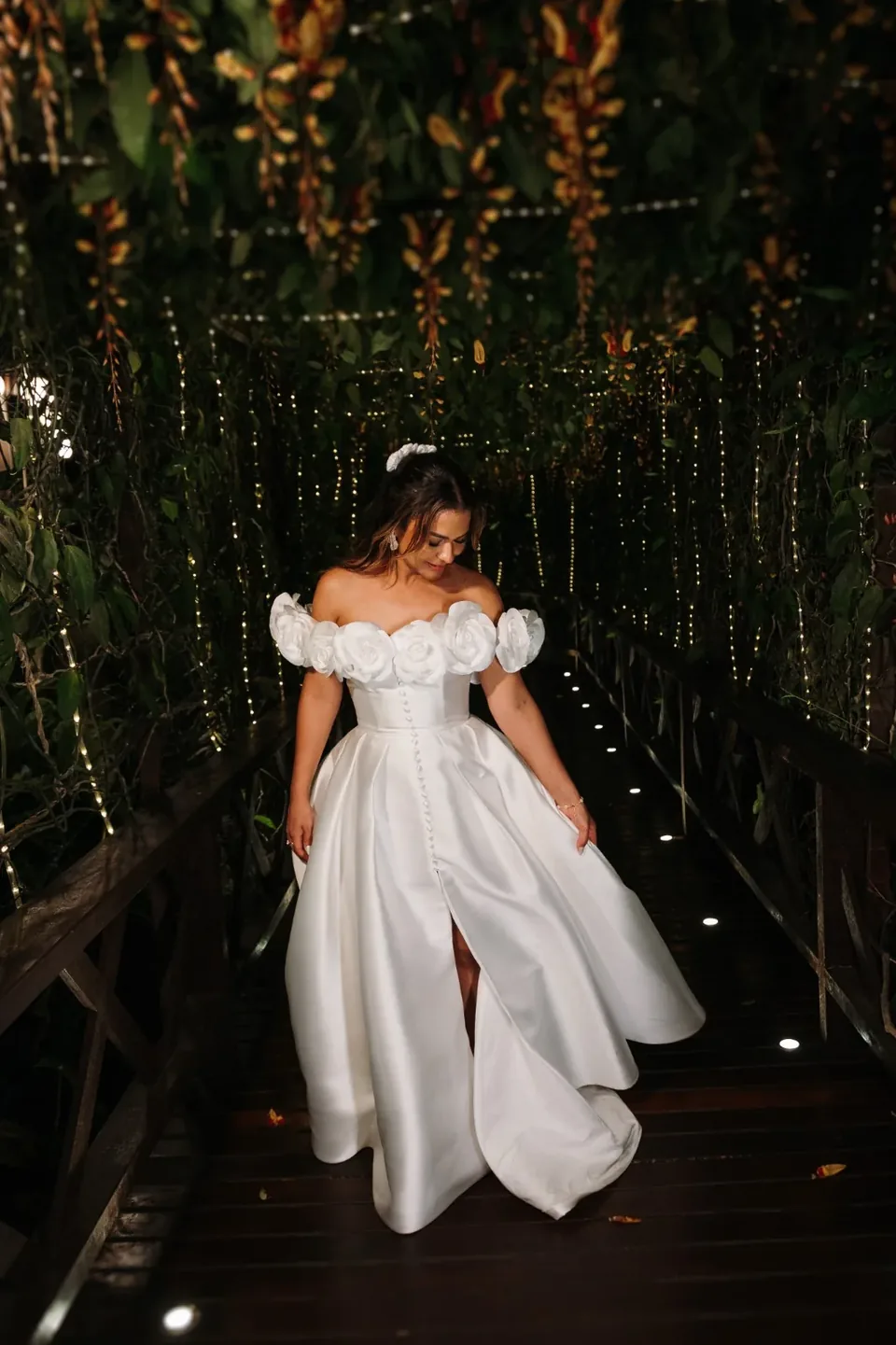 Noiva de vestido branco na passarela de madeira iluminada por luzes suspensas