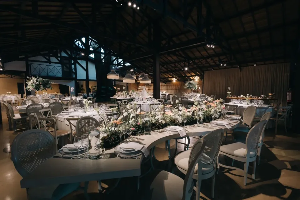 Mesa longa com arranjos de flores claras e cadeiras de palhinha no salão